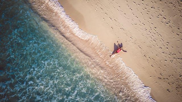An aerial view of a woman in a one-piece bathing suit lying on a beach.
